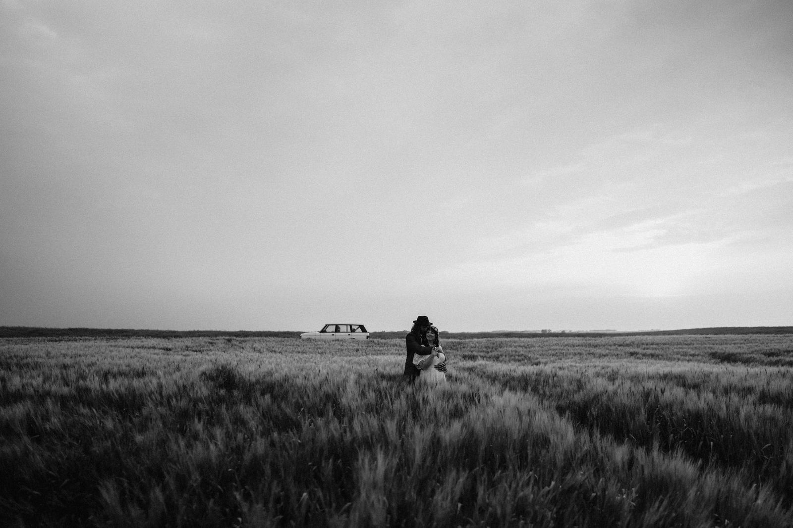 A couple alone in a vast field of tall grass, a car far behind them, black and white